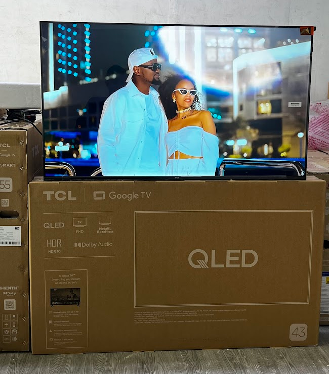 TV displaying a couple dressed in white standing on a glass railing against a nighttime city backdrop, atop a TCL Google TV box package in a showroom/retail setting.