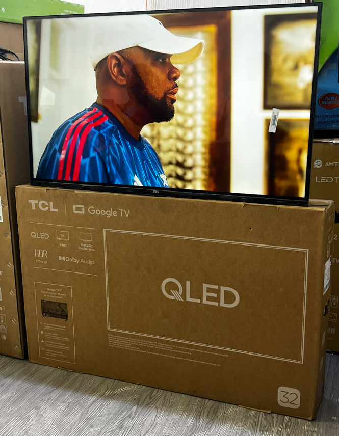 b – TCL East Africa Kenya Television displaying a man in a white cap and blue shirt, resting on a cardboard TCL box in a store.