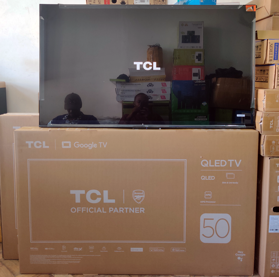 50t6c – TCL East Africa Kenya Flat-screen TCL Google TV 50 on its box in a warehouse; reflection of a person taking the photo is visible on screen.